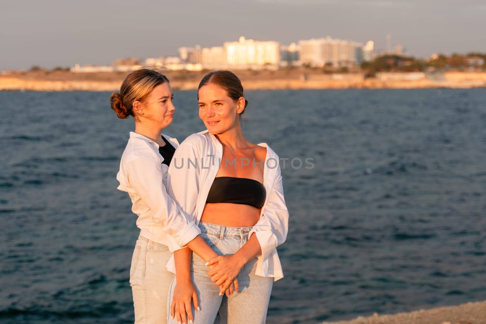 Two women stand close together on a beach, looking at each other. One wears a white shirt and blue jeans, the other a black bandeau top and white shirt. The sea and a city skyline are in the background.