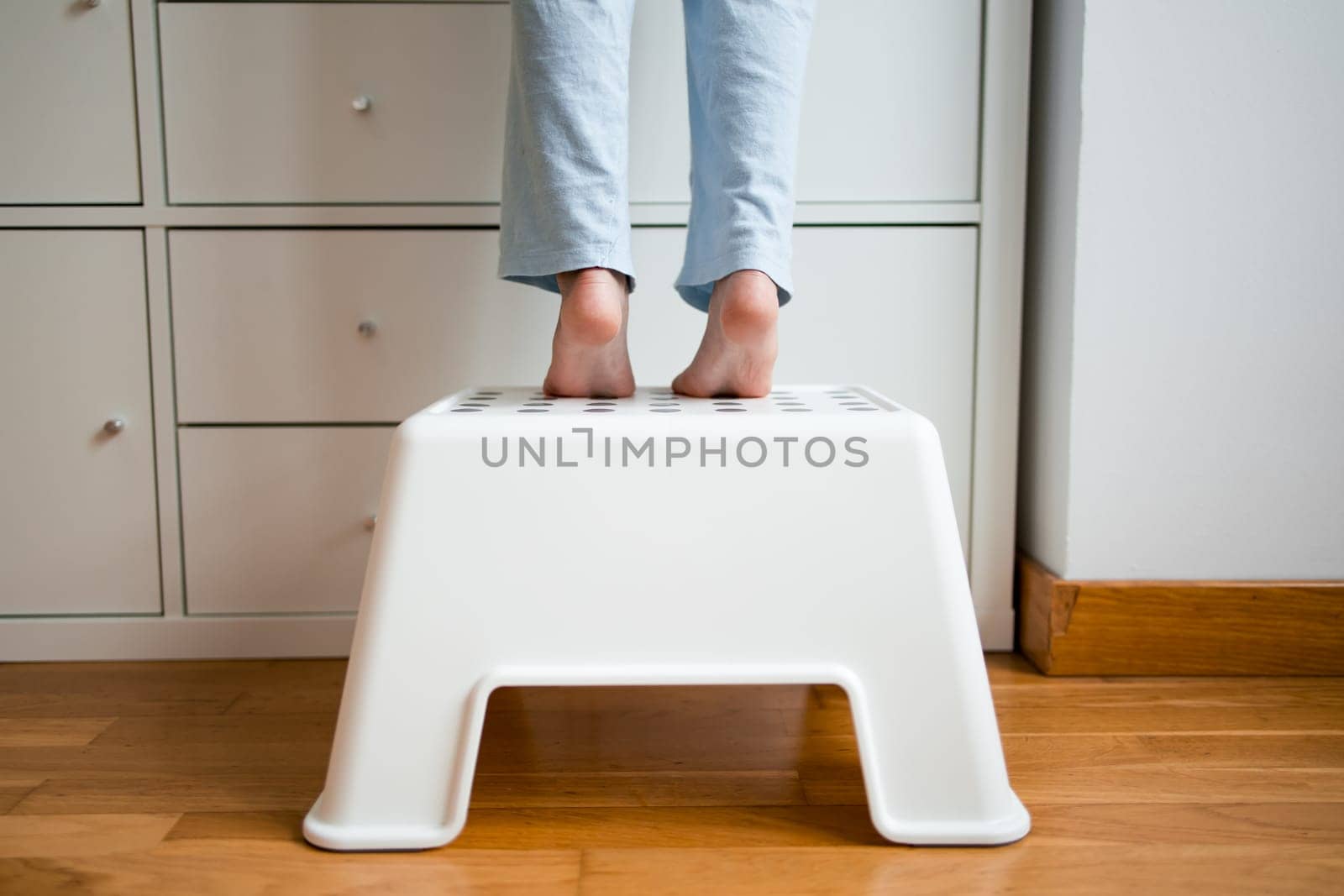Caucasian child standing on white step stool reaching high cabinet.