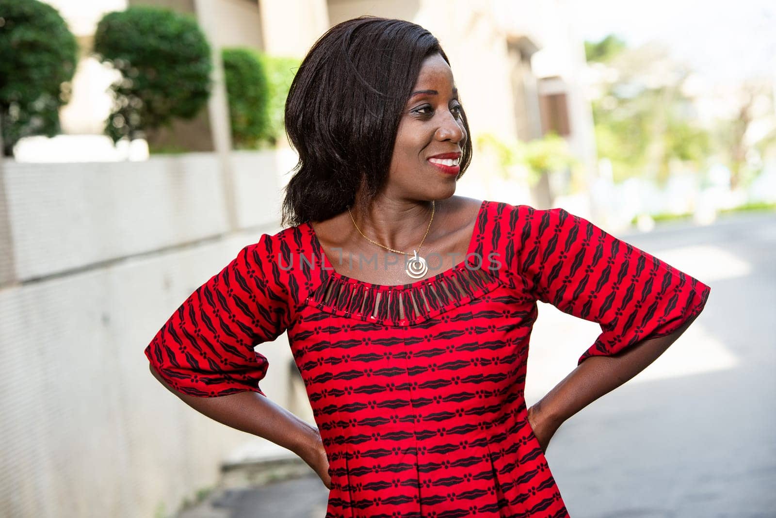Stylish African woman in fashion, wearing a red loincloth and standing on a street, hands on hips looking at her side.