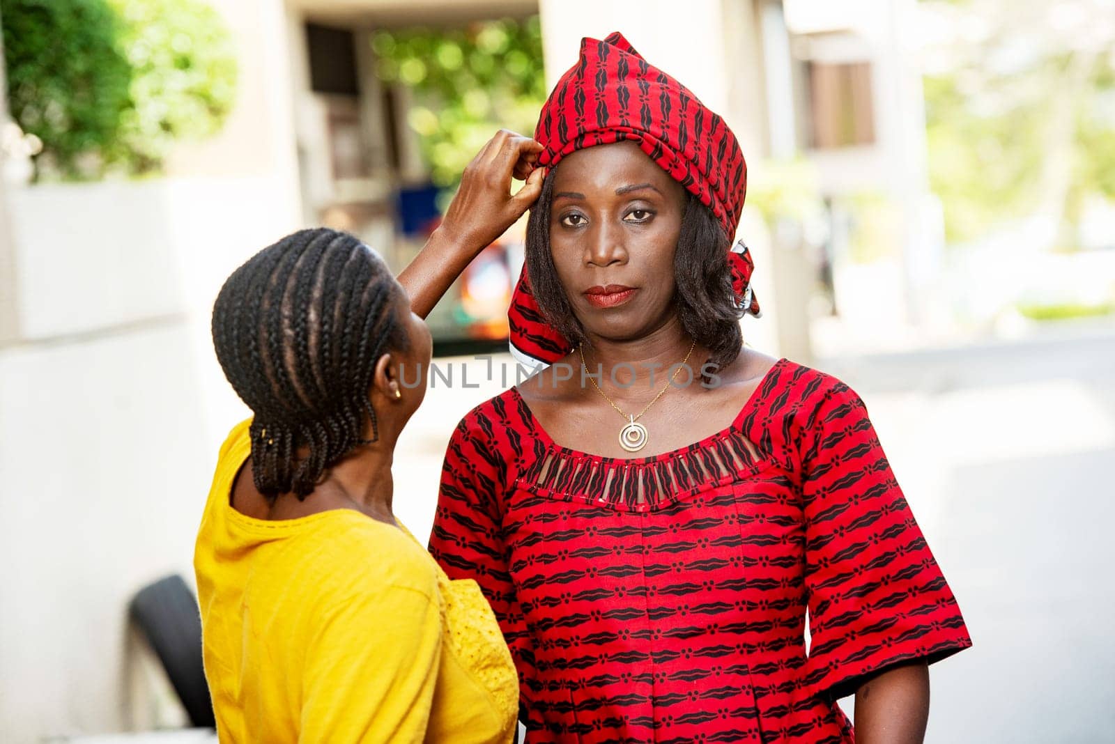 mature african woman standing outdoors looking at the camera while her sister adjusting her headscarf.