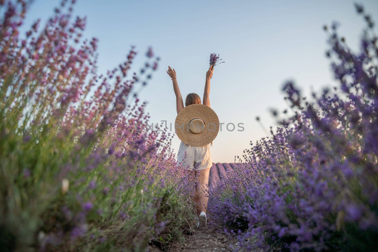 A woman is walking through a field of purple flowers with a straw hat on.