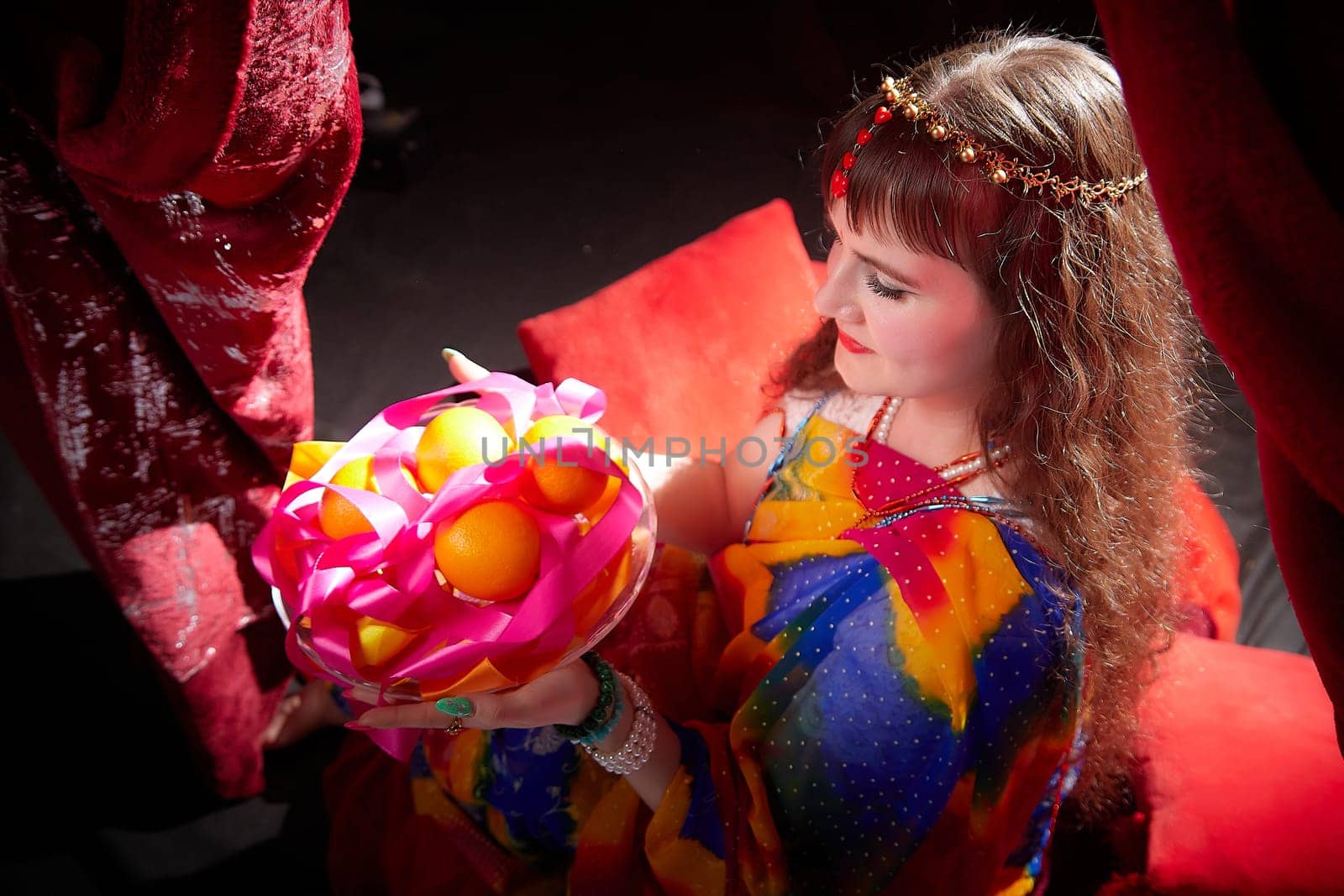 A mature woman with blonde hair showcases a beautiful sari while holding an elaborate fruit arrangement, surrounded by rich textiles and colors.