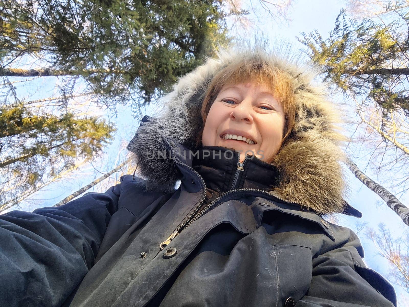 A mature blonde woman smiles brightly while taking a selfie in a snowy forest surrounded by tall trees.