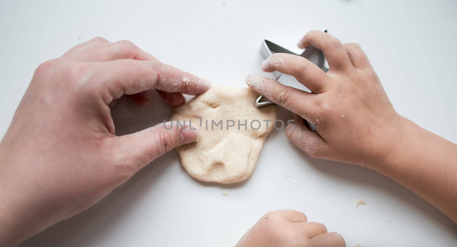 Child and adult hands crafting dough with cookie cutter on white surface.
