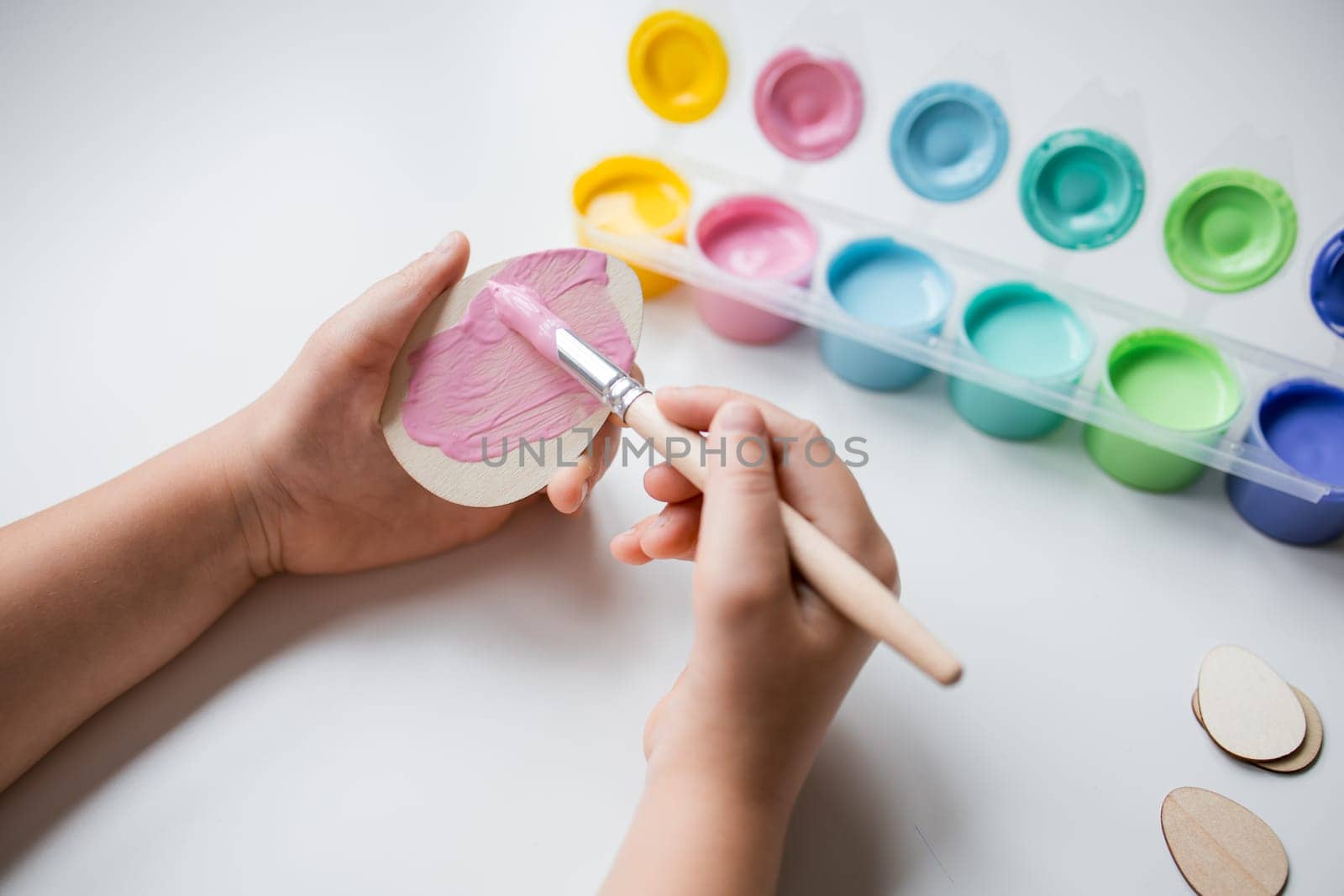 Child painting Easter wooden egg using pink acrylic paint on white surface.