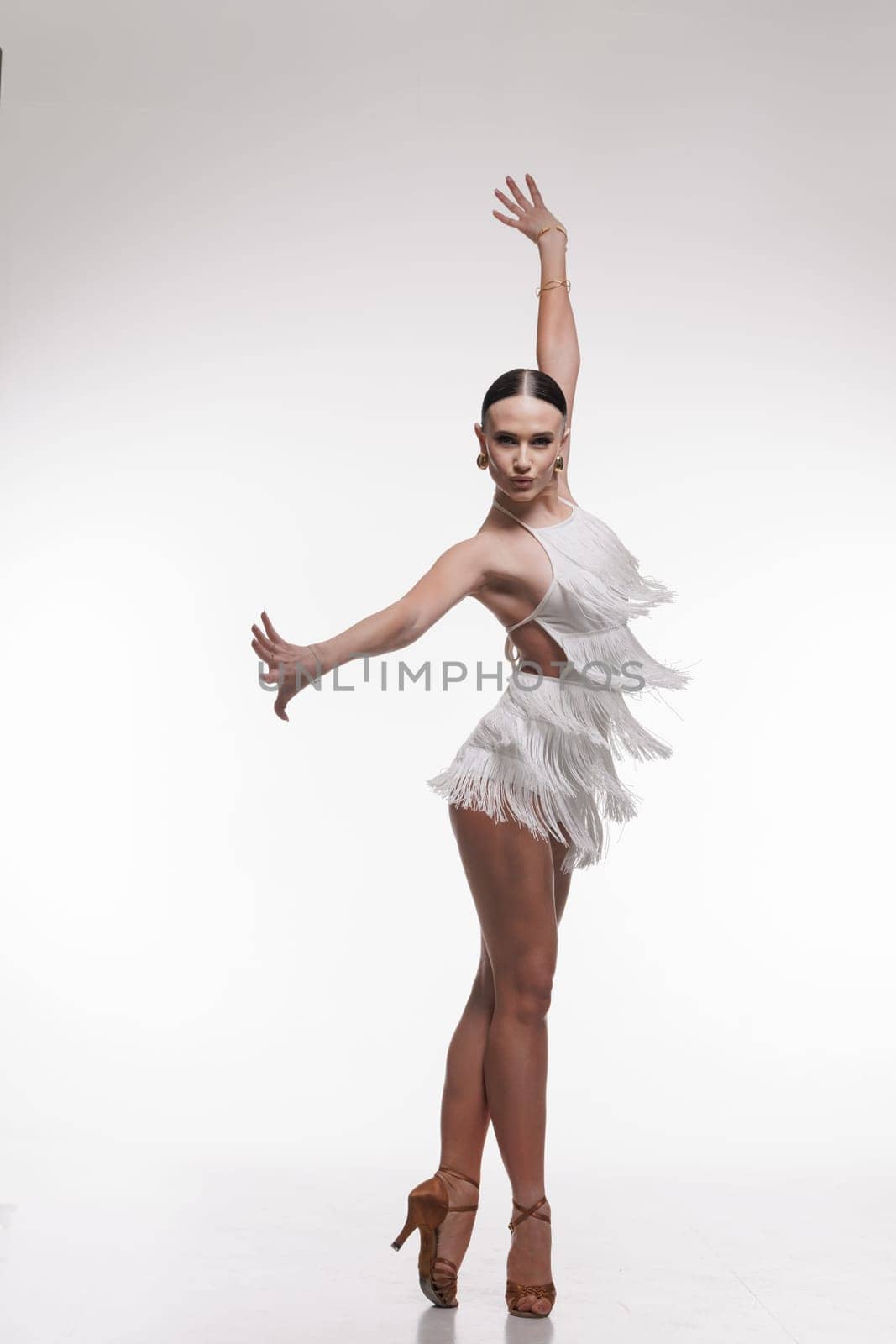 Elegant female dancer in white fringe outfit performing Latin dance pose on white background.