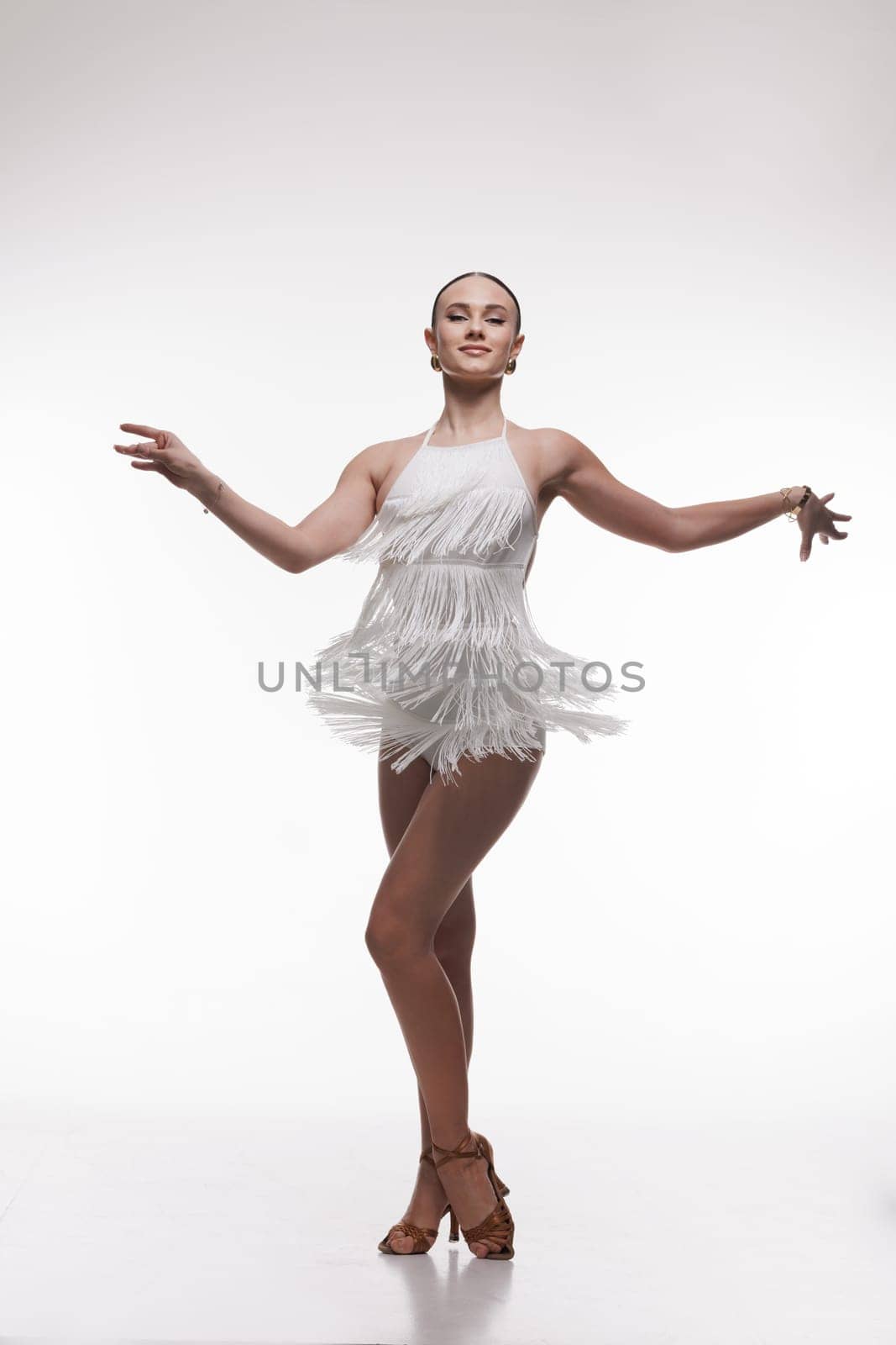 Elegant female dancer in white fringe outfit performing Latin dance pose on white background.