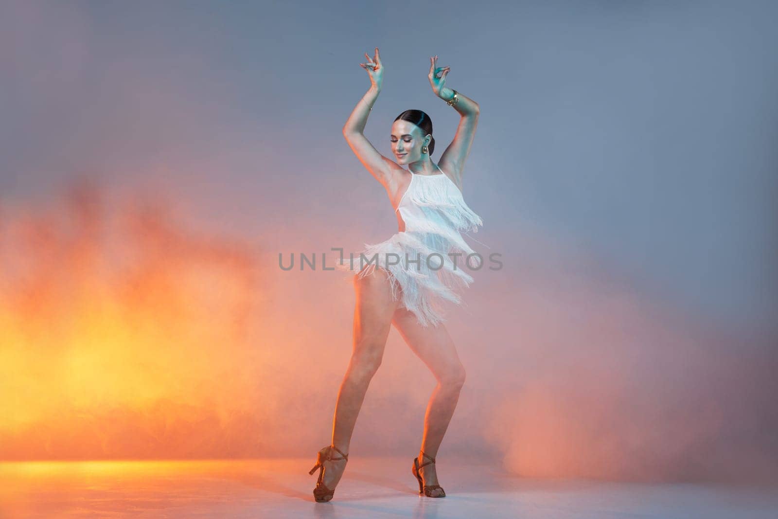 Elegant female Latin dancer in a white fringed dress performing a dynamic move with grace and confidence in artistic lighting.
