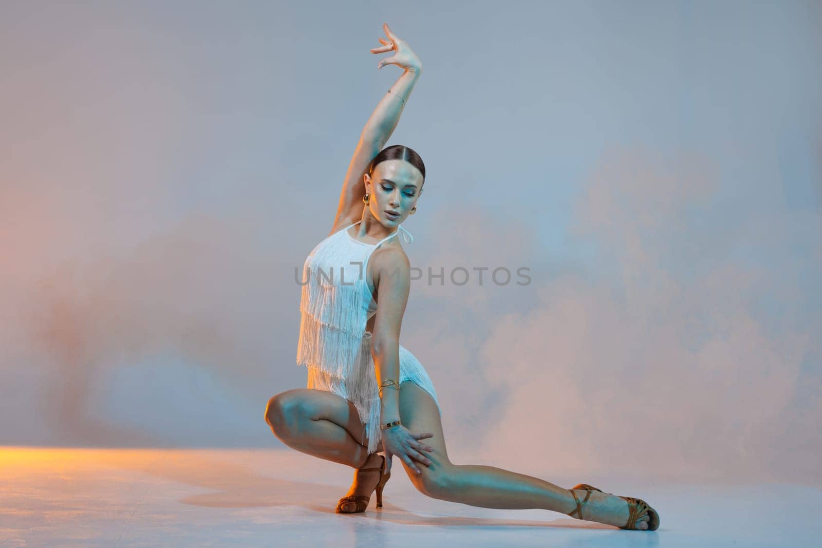 Elegant female Latin dancer in a white fringed dress performing a dynamic move with grace and confidence in artistic lighting.