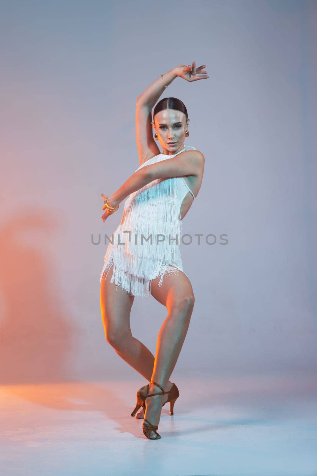 Elegant female Latin dancer in a white fringed dress performing a dynamic move with grace and confidence in artistic lighting.