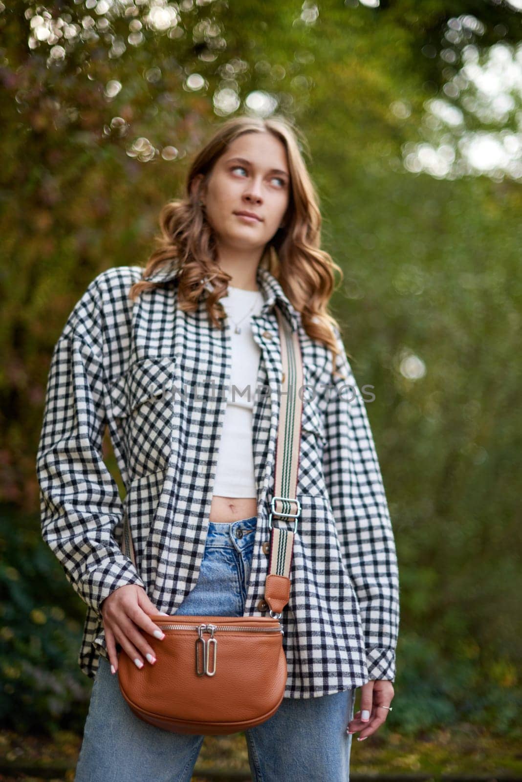 A young woman with wavy brown hair and blue eyes confidently poses outdoors in a lush autumn park. She wears a trendy black-and-white checkered shirt over a white crop top and classic blue jeans, accessorized with a tan leather crossbody bag with a striped strap. The soft natural lighting and blurred background of green foliage create a dreamy and cozy fall atmosphere. This fashionable portrait is ideal for lifestyle branding, autumn fashion campaigns, social media promotions, and advertising seasonal collections.