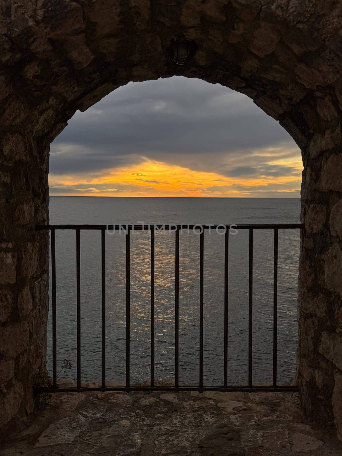 A rustic stone archway frames a view of calm blue waters and a golden sunset, with a black iron railing in the foreground. High quality photo. High quality photo
