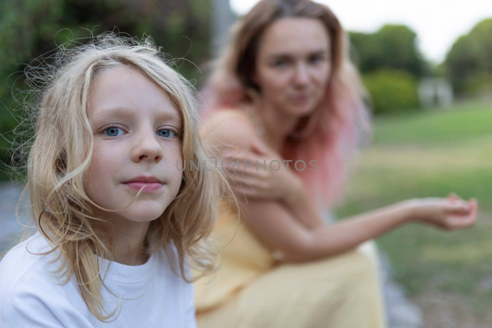 Young blonde child posing in a park with mother sitting in blurred background