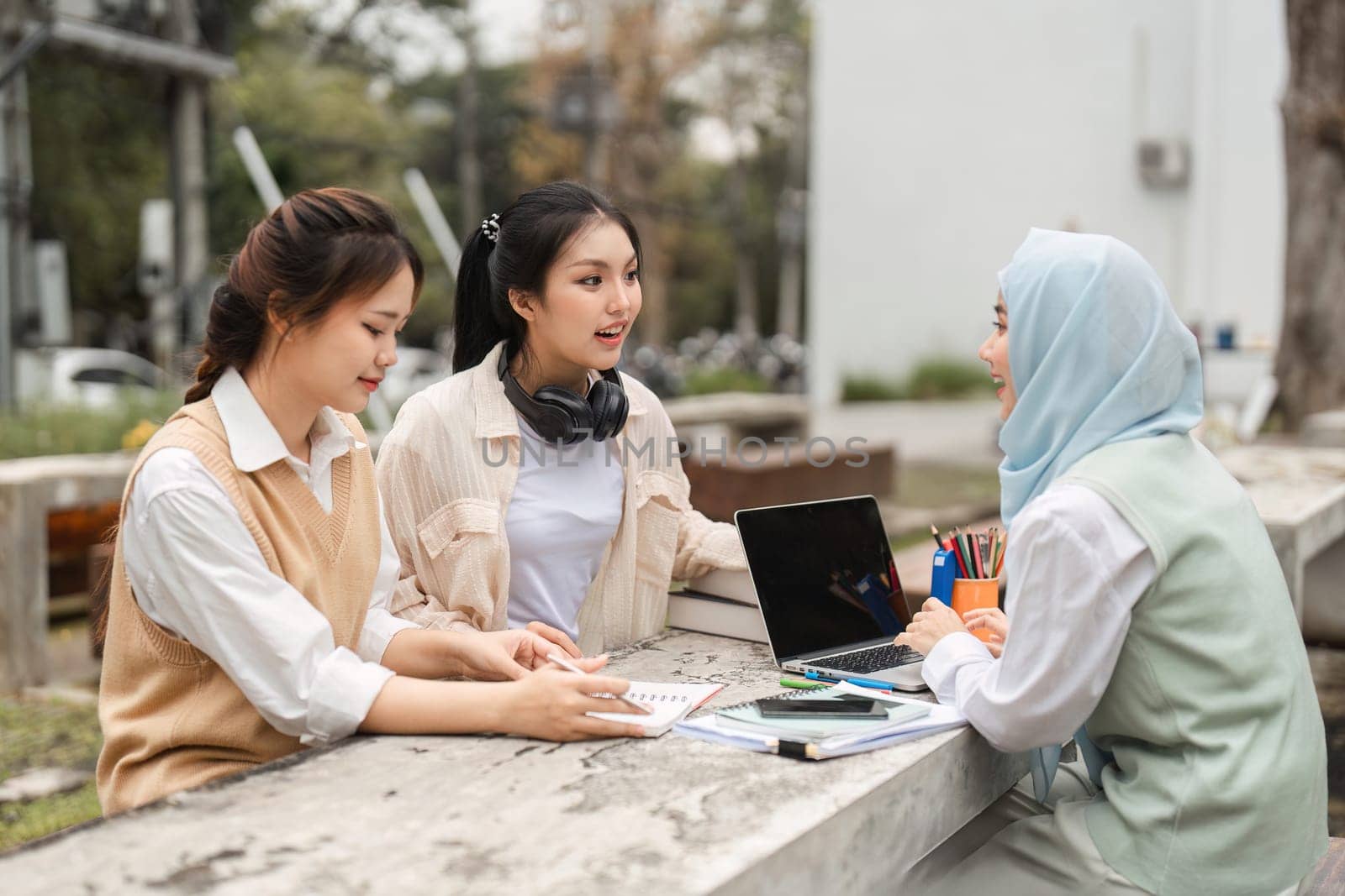 A group of multicultural women are engaged in a lively discussion, sharing ideas with a laptop and notebooks while sitting outdoors in a university environment.