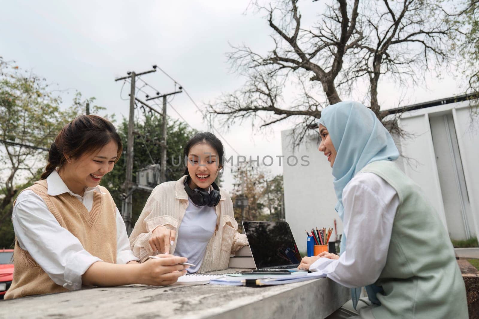 A diverse group of university students joyfully collaborating on academic tasks outdoors, using a laptop and sharing notes and ideas.