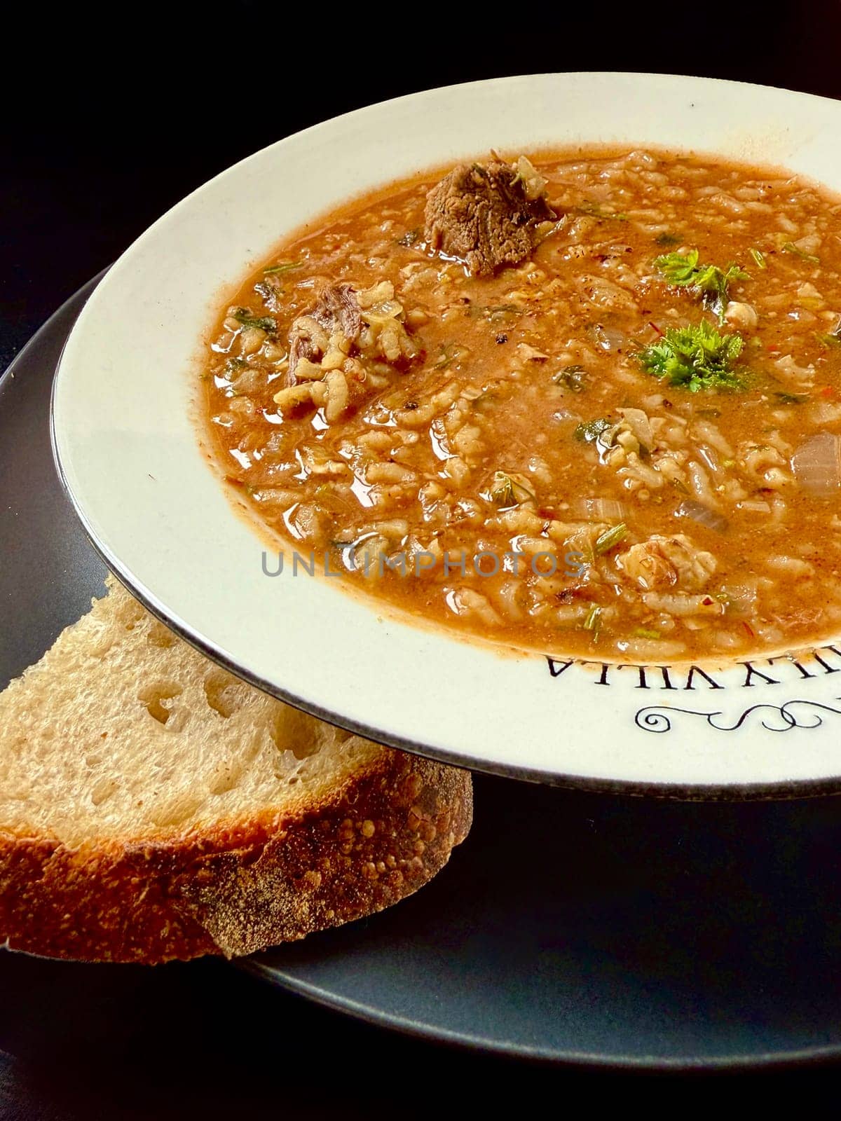Georgian soup. Kharcho with rice, tomatoes, beef, garlic and herbs in white plate on black table Traditional Caucasian food spicy soup with lamb, tkemali and rice. Man showing pieces of meat with spoon.