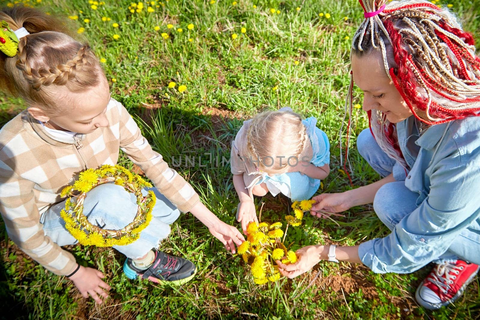 A mother and her two children gather dandelions and create colorful flower crowns together in a bright, green meadow.