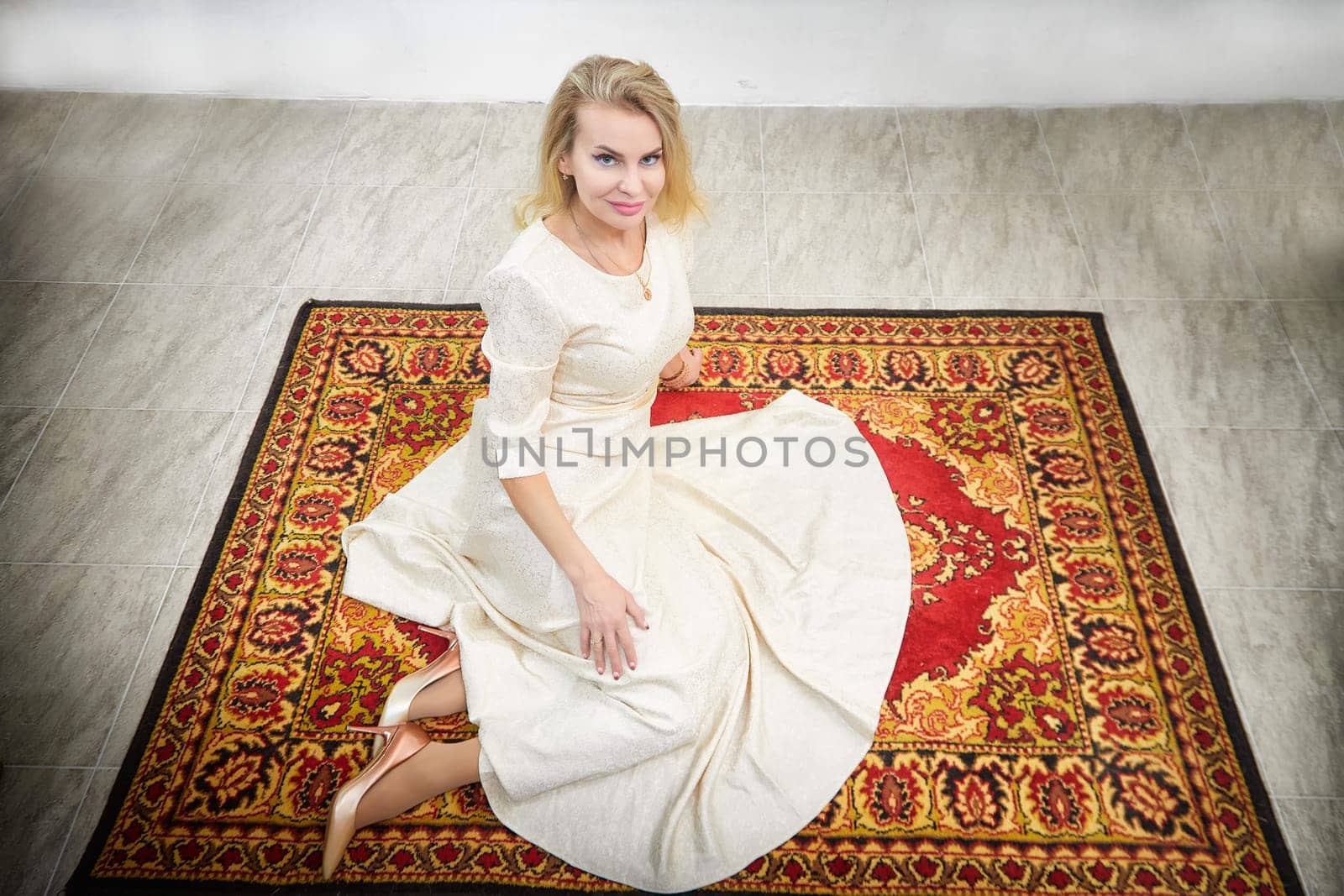 This middle-aged blonde blogger models in a stylish dress while posing on a decorative rug inside her home.