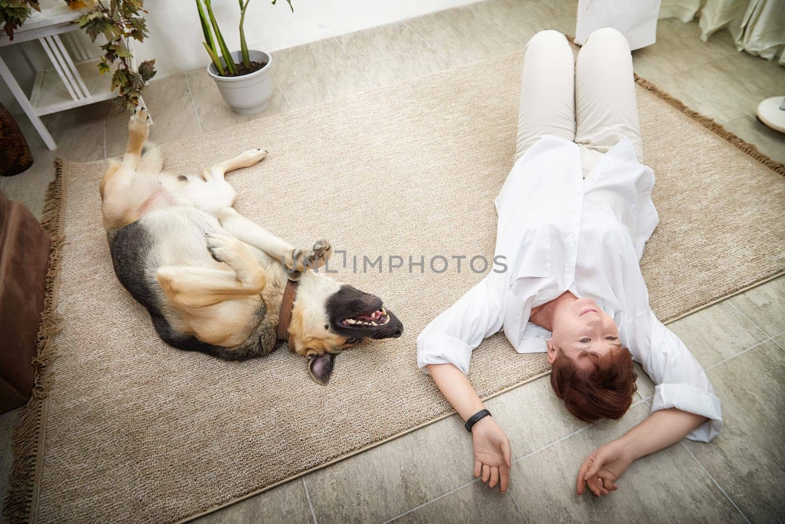Middle-aged woman lies on the floor next to her happy German Shepherd in a warm, inviting room.