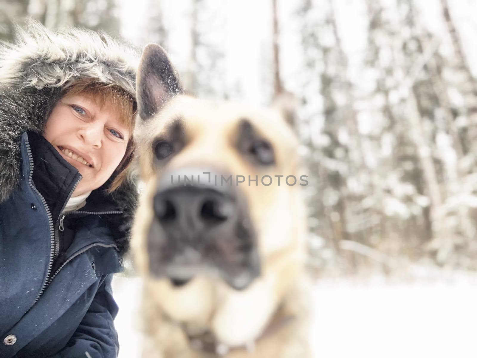 Smiling woman poses with her dog in a snowy forest during winter, surrounded by tall trees.