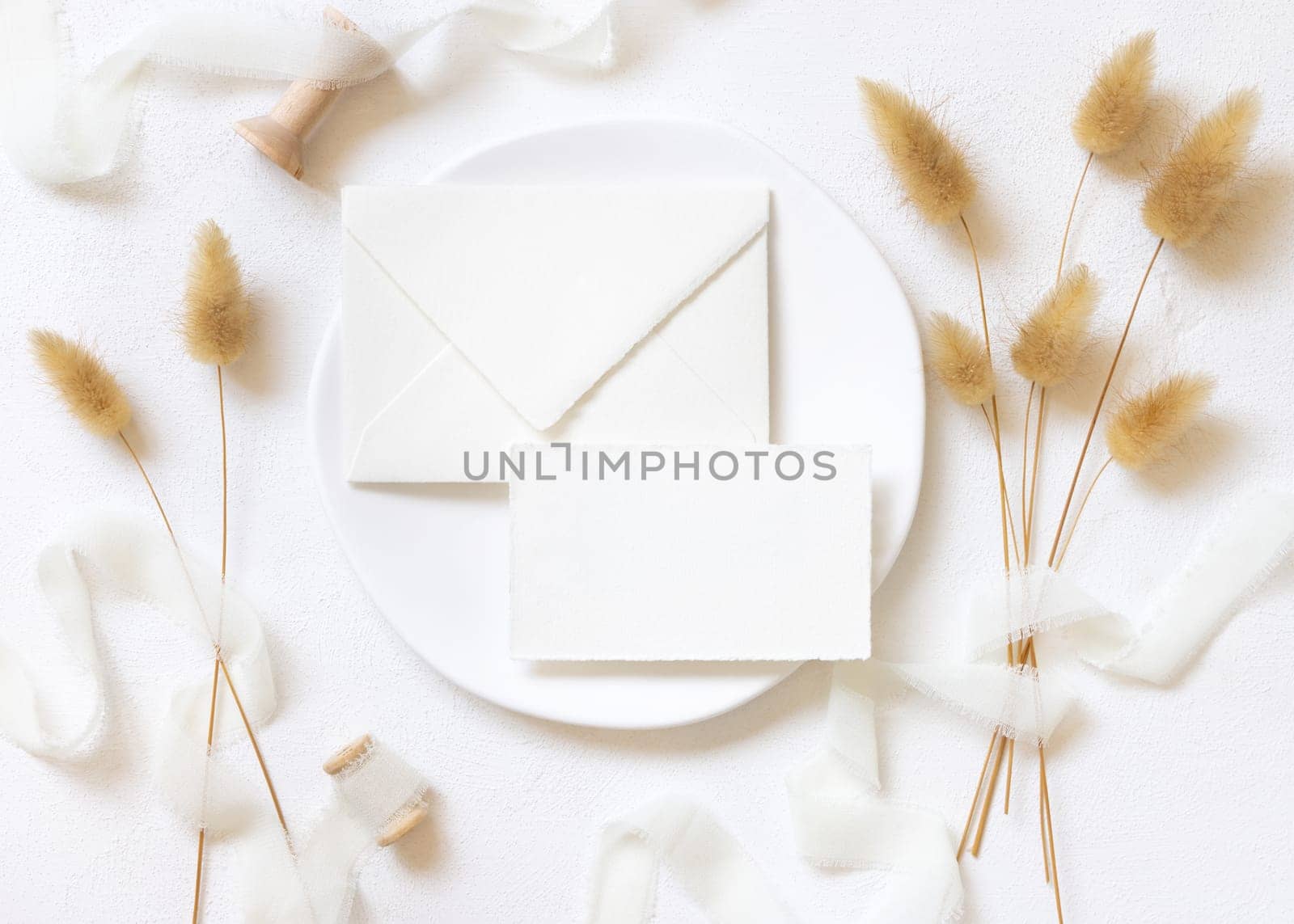 Blank card and envelope on white plate near curled silk ribbons and dried hare's tail grass topview, mockup. Pastel romantic flatlay. Bohemian or close to nature cards for party design or wedding stationery
