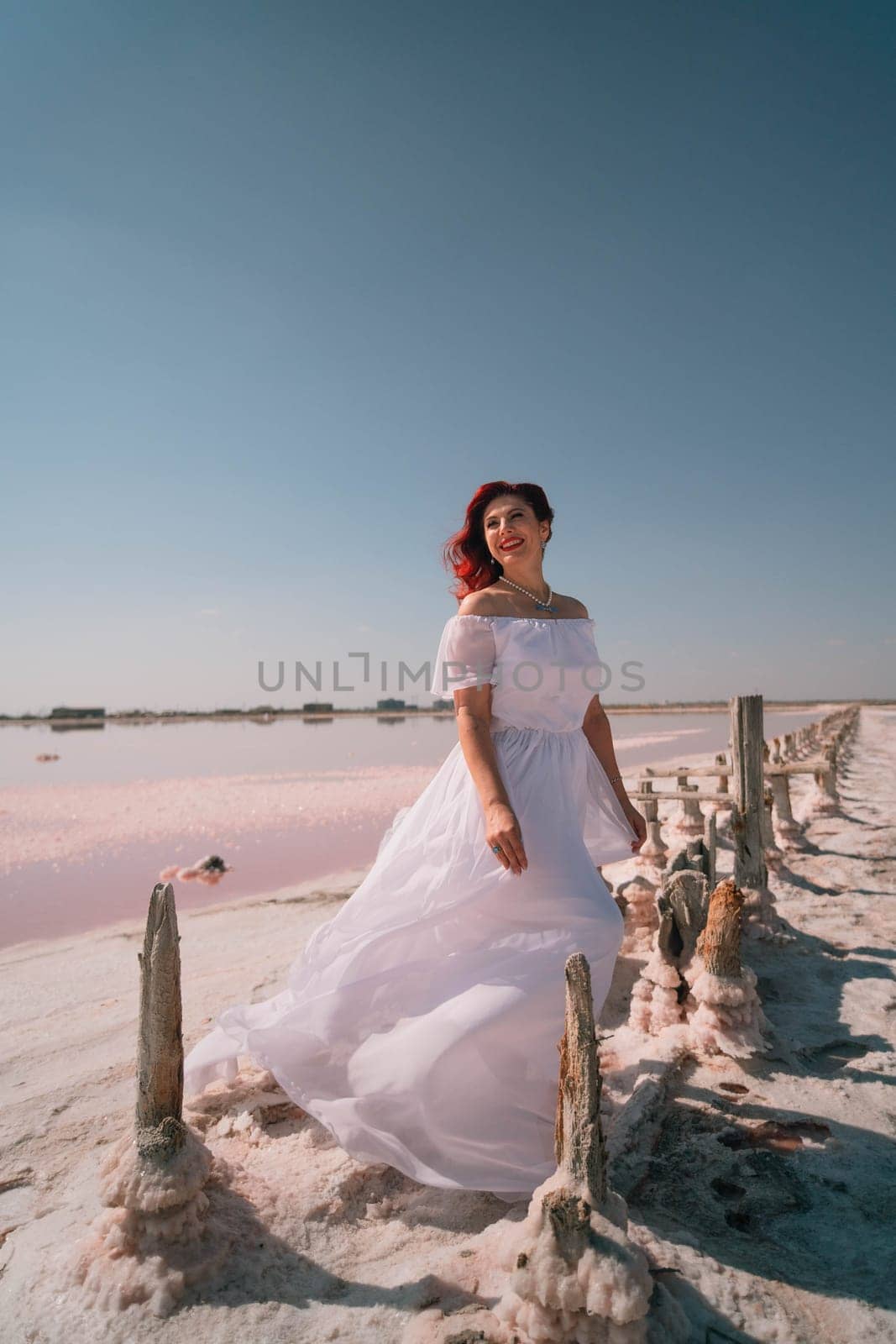 A woman in a white wedding dress stands on a wooden fence, overlooking a pink salt lake. The sun is shining and the sky is blue.