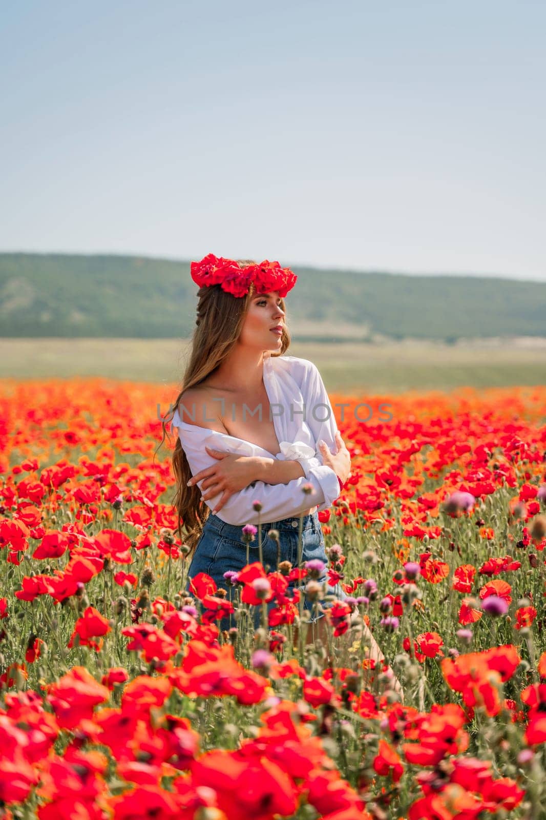 Happy woman in a poppy field in a white shirt and denim skirt with a wreath of poppies on her head posing and enjoying the poppy field