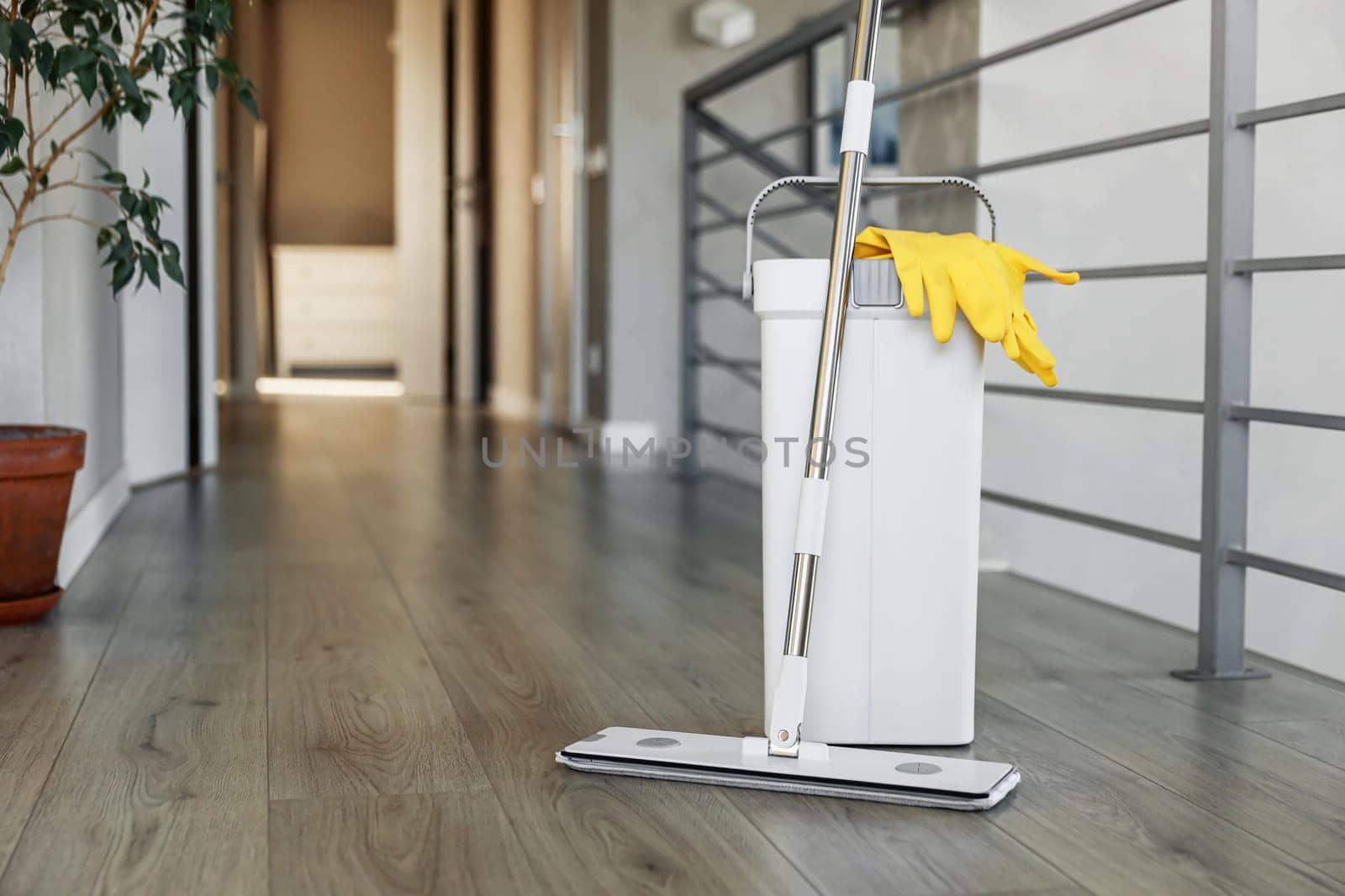 Cleaning supplies: mop, gloves, bucket with wringer sitting on wooden laminate floor in modern living room, wet equipment tools, housekeeping concept. Emphasizing home sanitation, cleanliness, hygiene