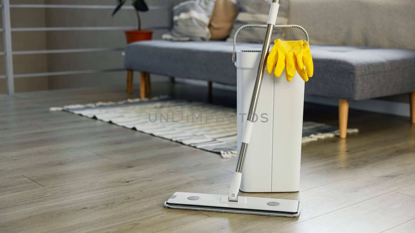Cleaning supplies: mop, gloves, bucket with wringer sitting on wooden laminate floor in modern living room, wet equipment tools, housekeeping concept. Emphasizing home sanitation, cleanliness, hygiene