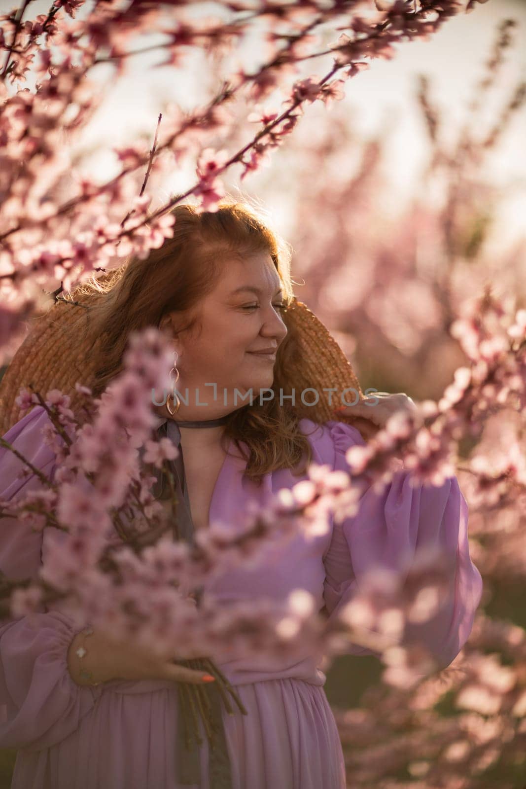 Woman blooming peach orchard. Against the backdrop of a picturesque peach orchard, a woman in a long pink dress and hat enjoys a peaceful walk in the park, surrounded by the beauty of nature