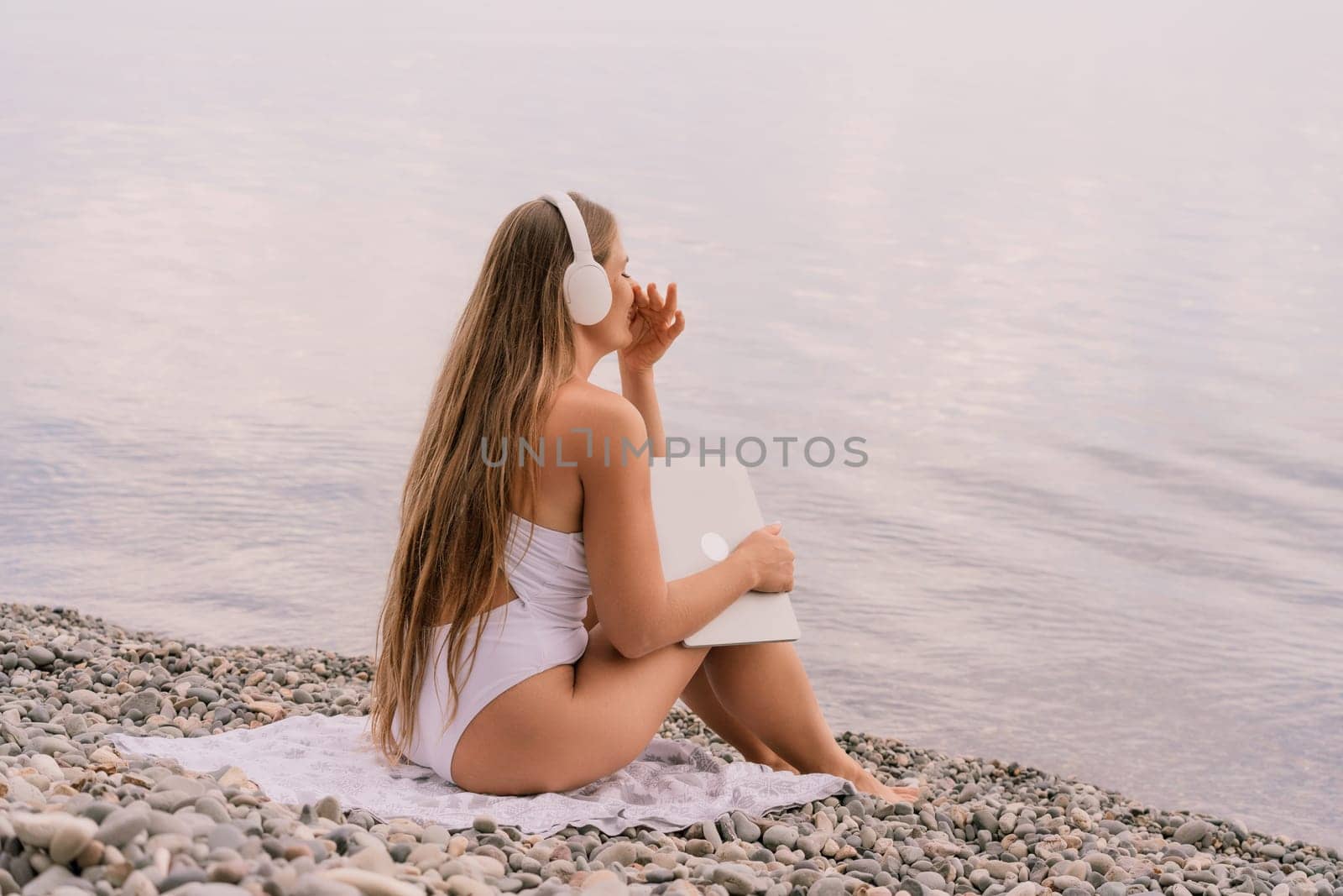 A woman is sitting on the beach with a laptop and headphones on. She is wearing a white bikini and she is enjoying her time by the water