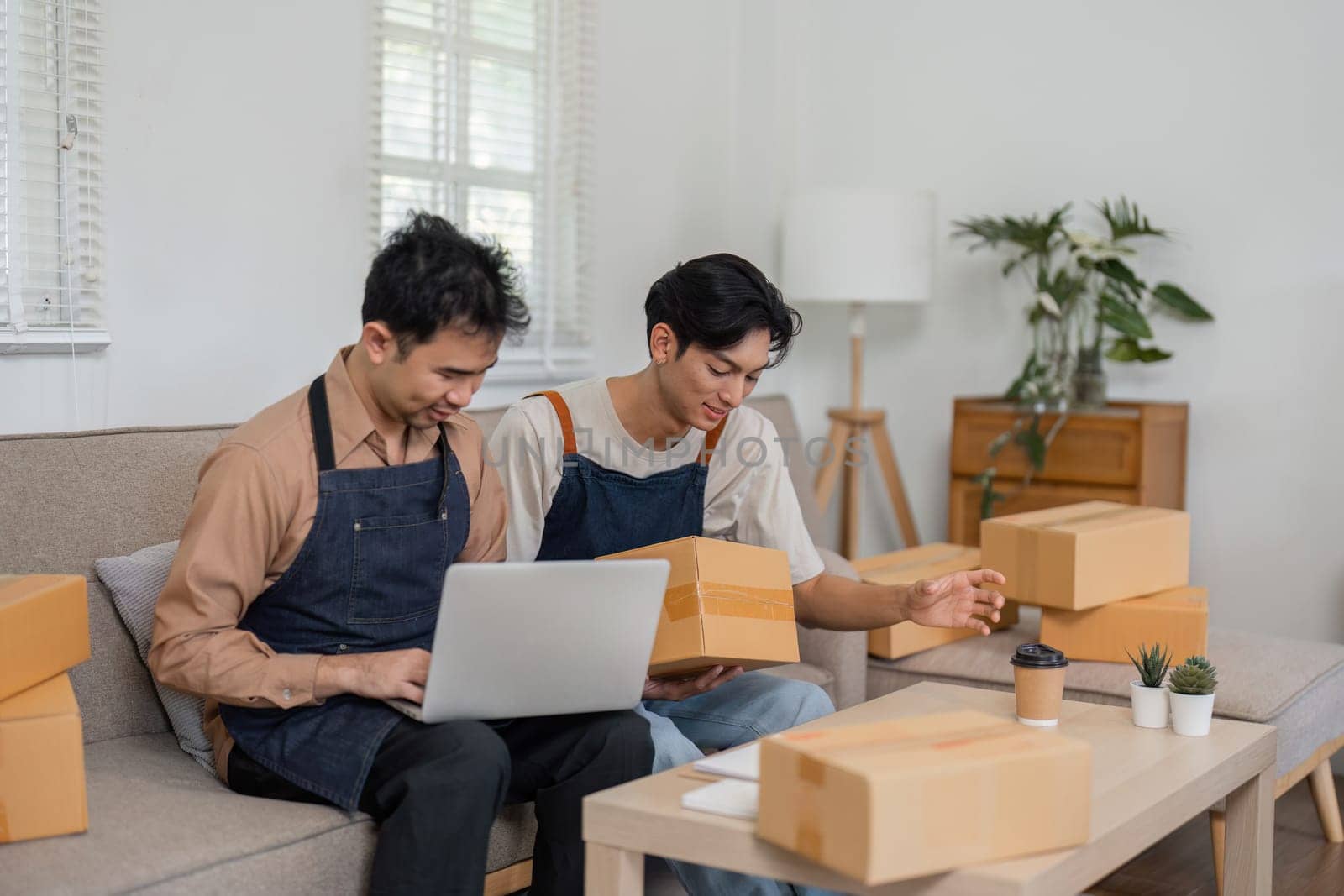 Two friendly social media marketers engaged in a discussion about product details, with one using a laptop and the other holding a package, in their modern home office filled with boxes.
