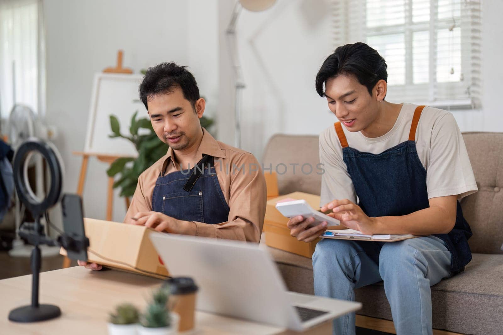 Two young Asian entrepreneurs reviewing product information on a smartphone while packaging items in their cozy home office for their e-commerce business.