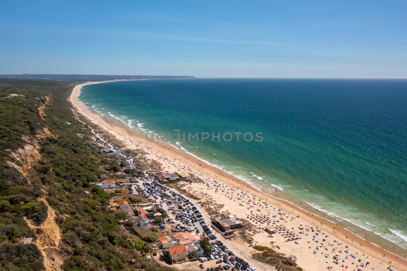 Aerial drone view showcases vibrant, crowded Fonte de Telha beach at Costa de Caparica, Portugal, with its golden sands, turquoise waters, and stunning seascape set against backdrop of blue sky