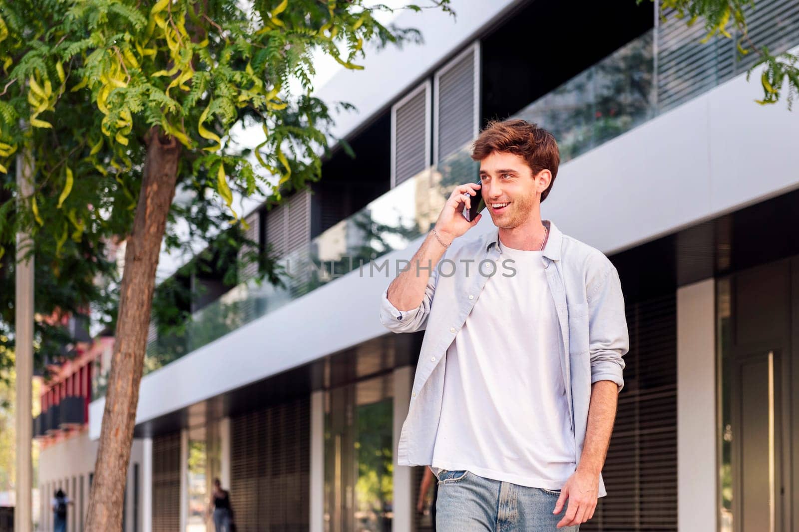 young man smiling happy while walking down the street in a good mood talking on mobile phone, concept of technology of communication and urban lifestyle, copyspace for text