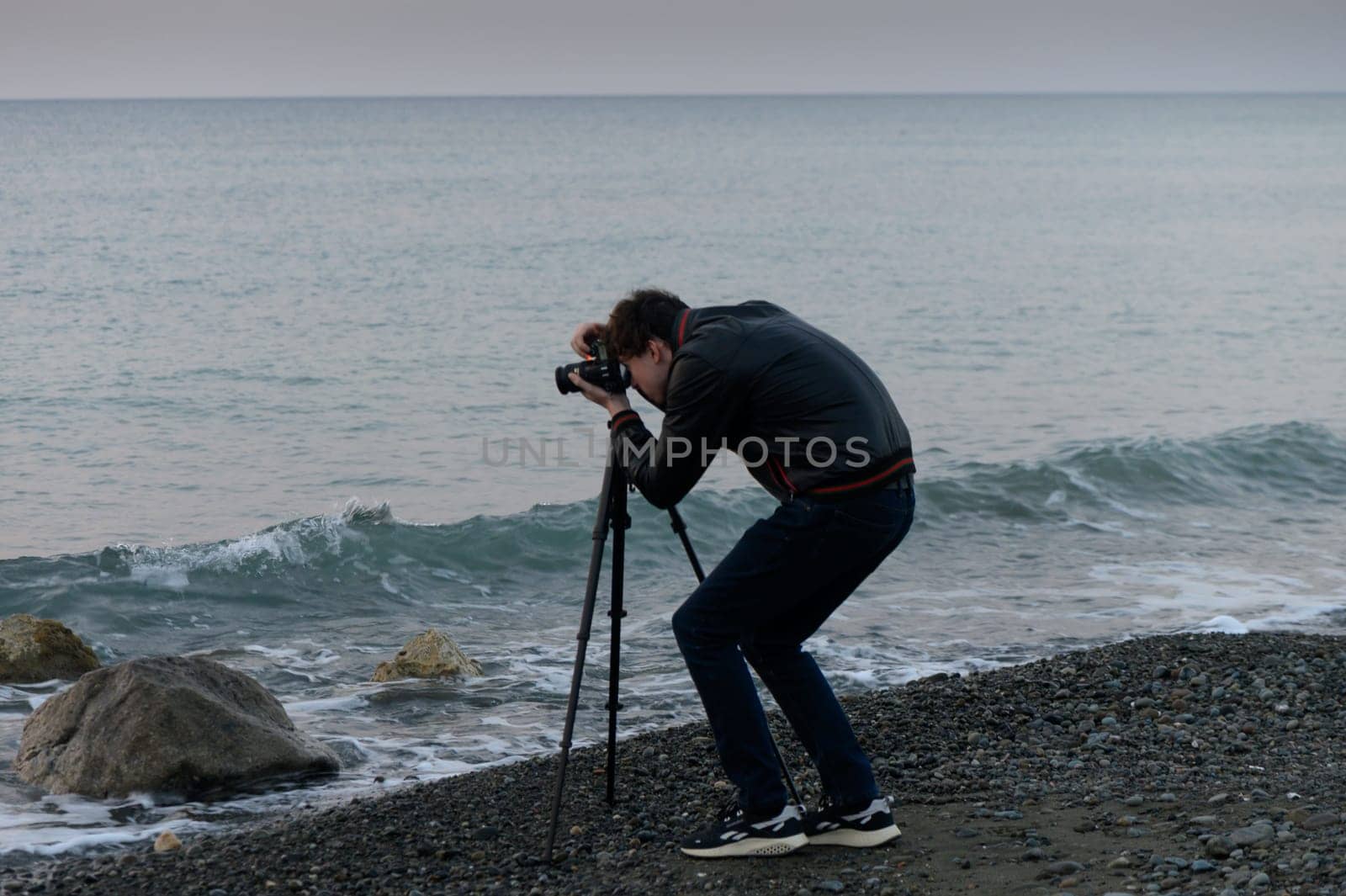 A photographer bends over his camera, skillfully framing the serene ocean waves at dusk near rocky shores.