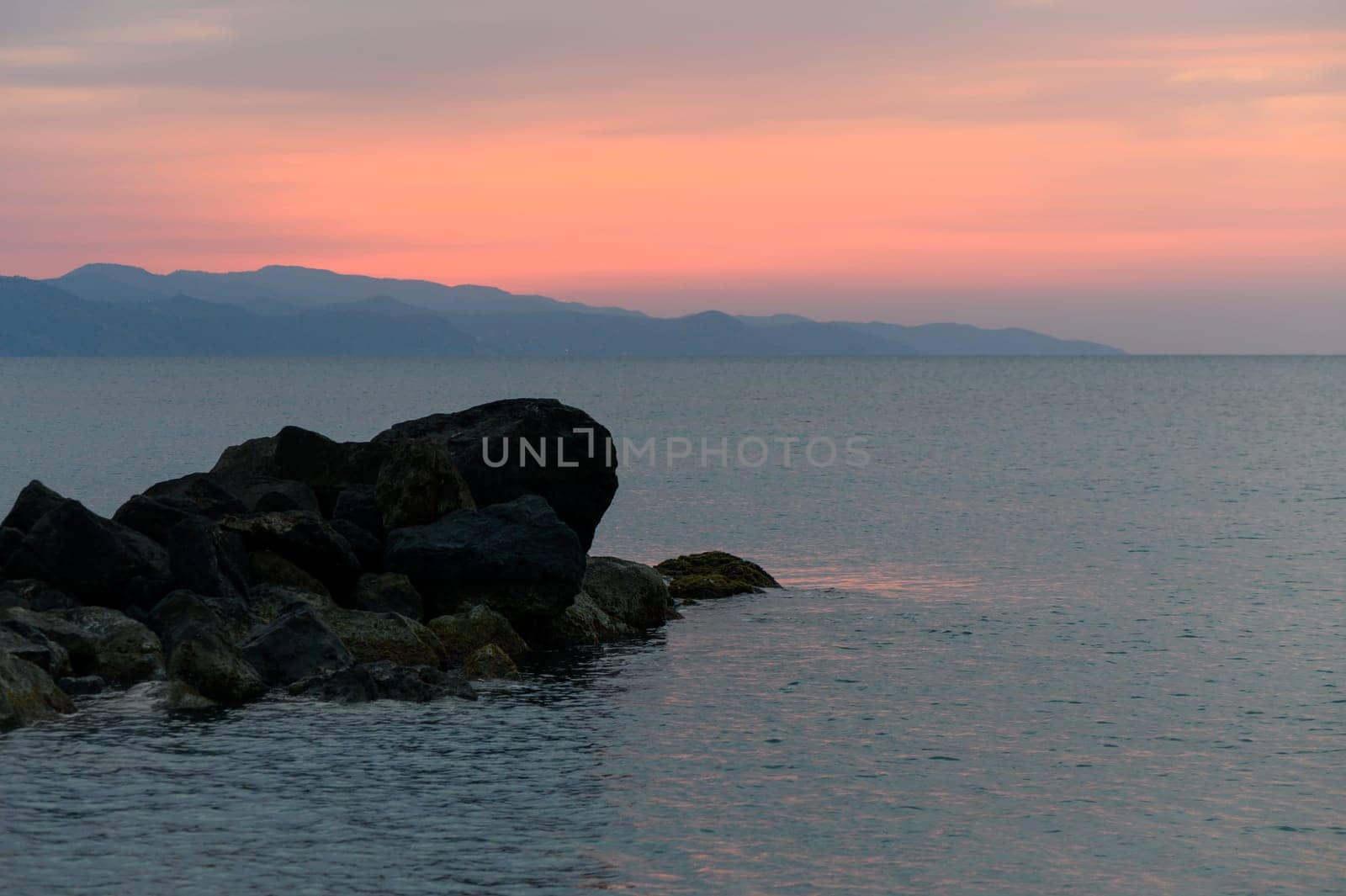 Calm waters reflect soft hues of orange and pink as waves gently lap against the rocks at dusk