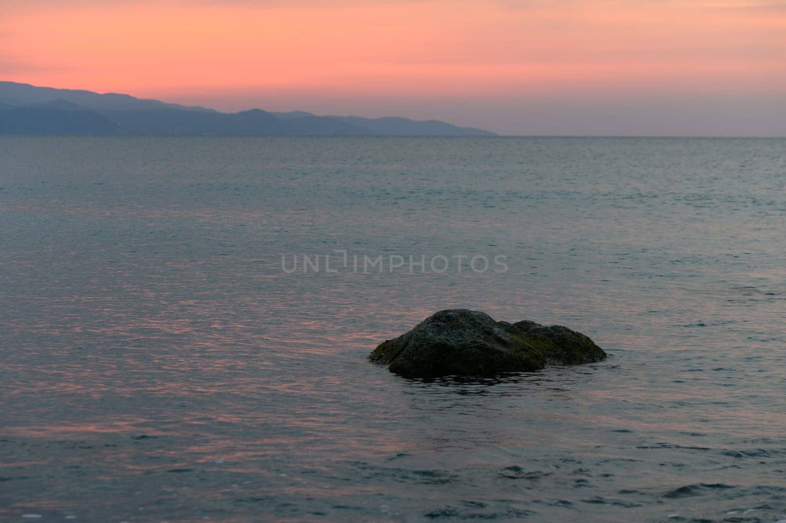 A serene view of calm waters reflecting the soft hues of sunset, with a solitary rock and distant mountains.