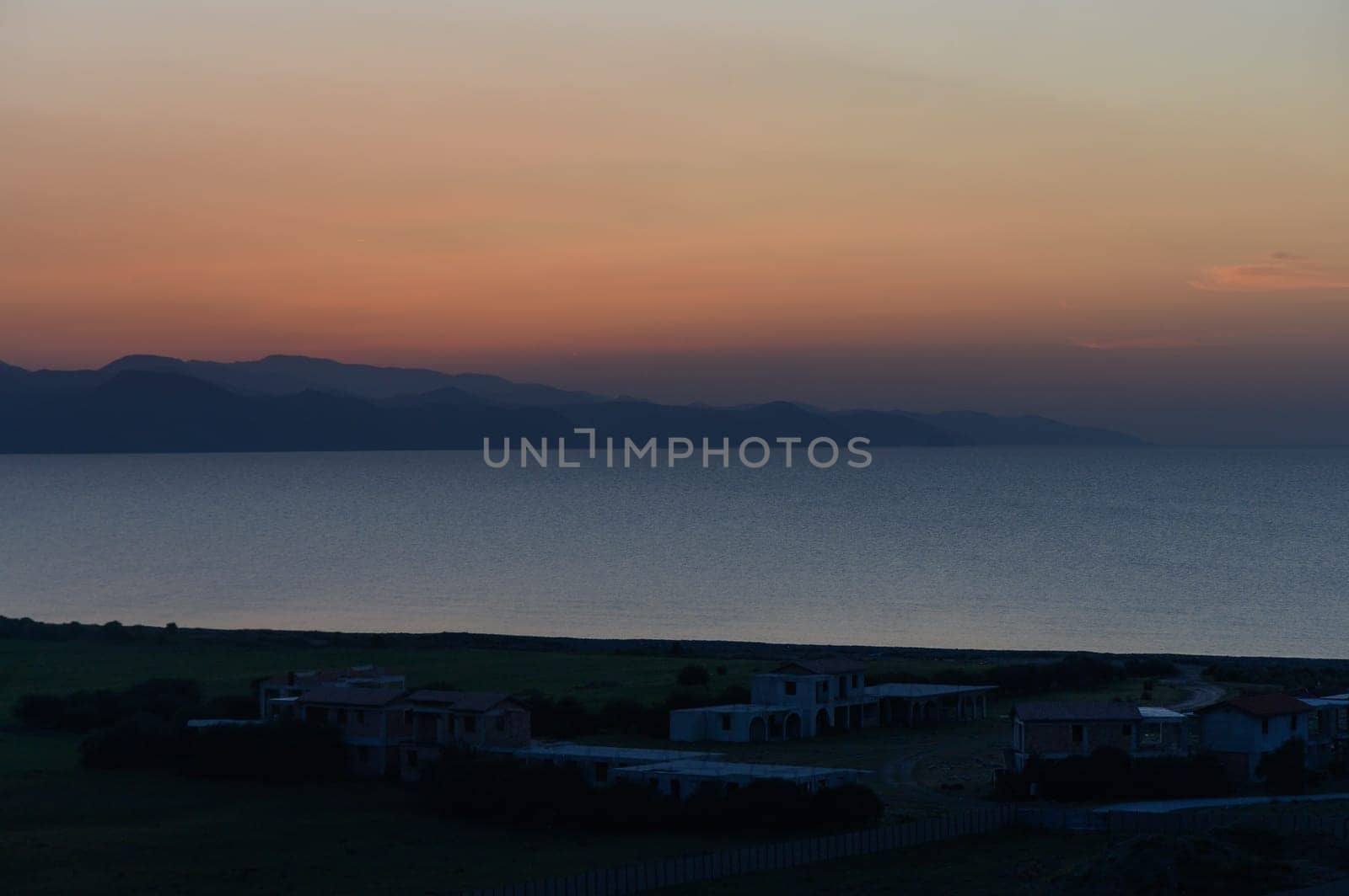 Soft hues of orange and purple illuminate the quiet shoreline as nightfall approaches over serene waters and hills.