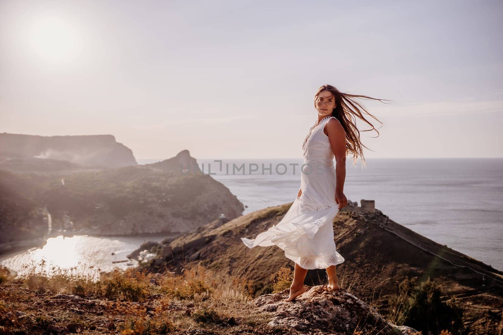 A woman in a white dress stands on a hill overlooking the ocean. The scene is serene and peaceful, with the woman's dress billowing in the wind. The combination of the ocean