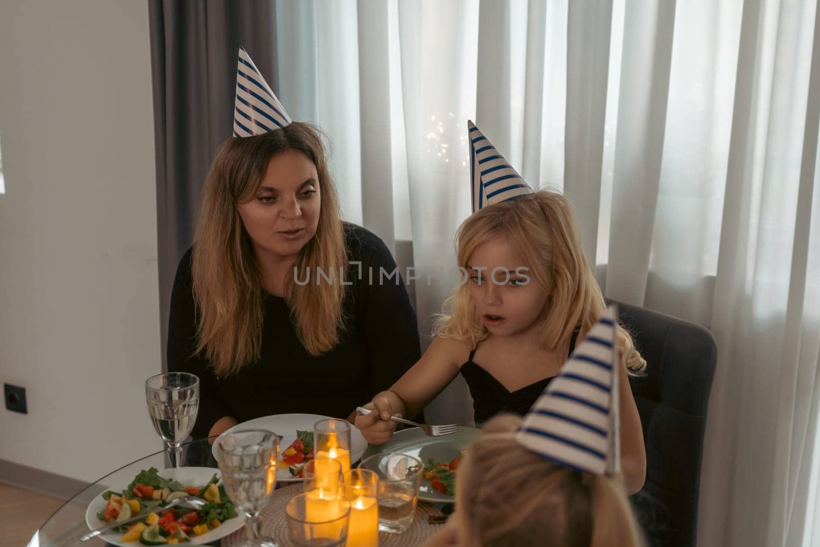 Woman and child wearing party hats sit at a table with candles, food, and drinks. Birthday celebration in progress.