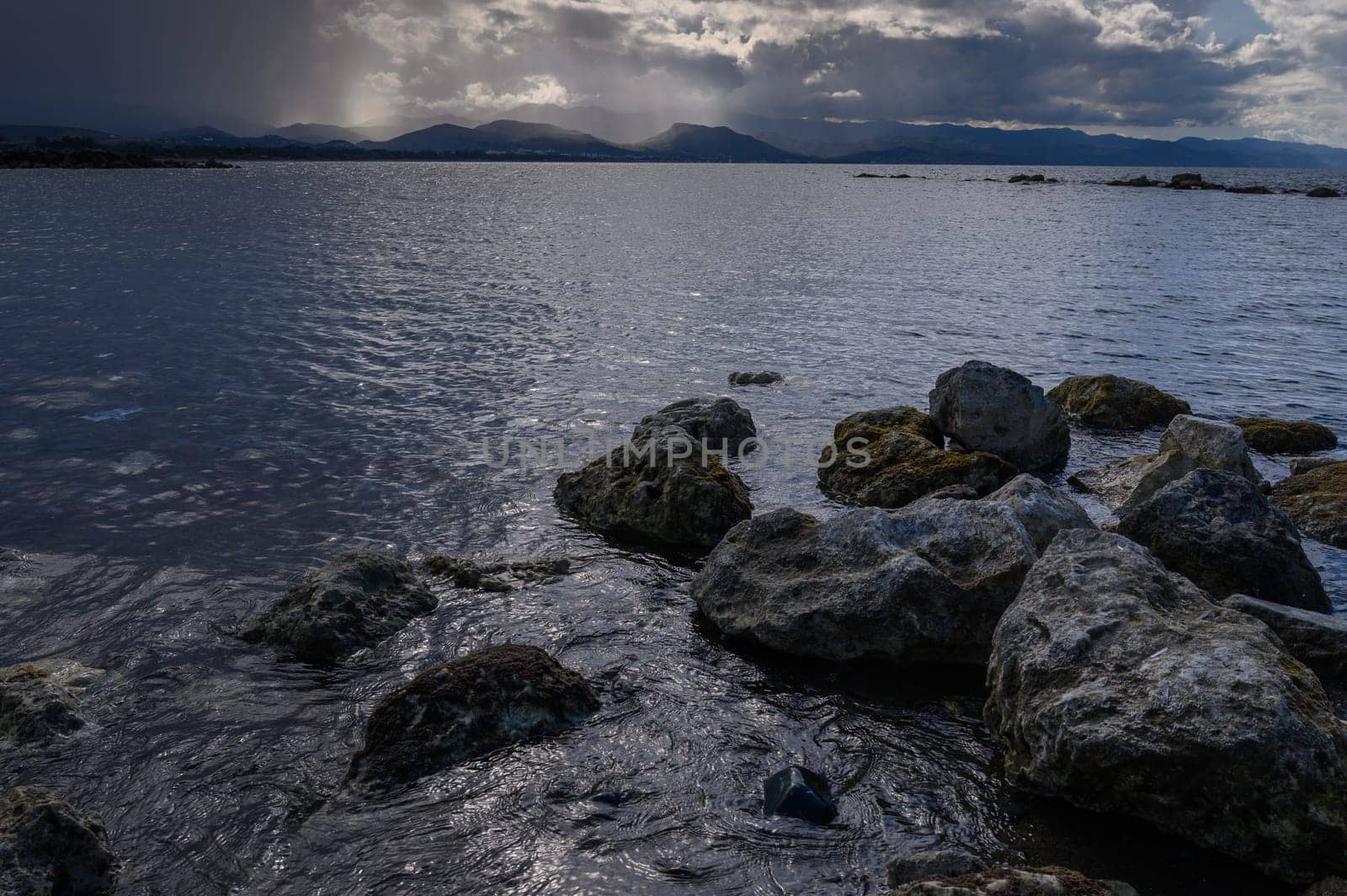 Dark clouds loom over the tranquil lake, casting shadows on the rocky shoreline as the sun begins to set.
