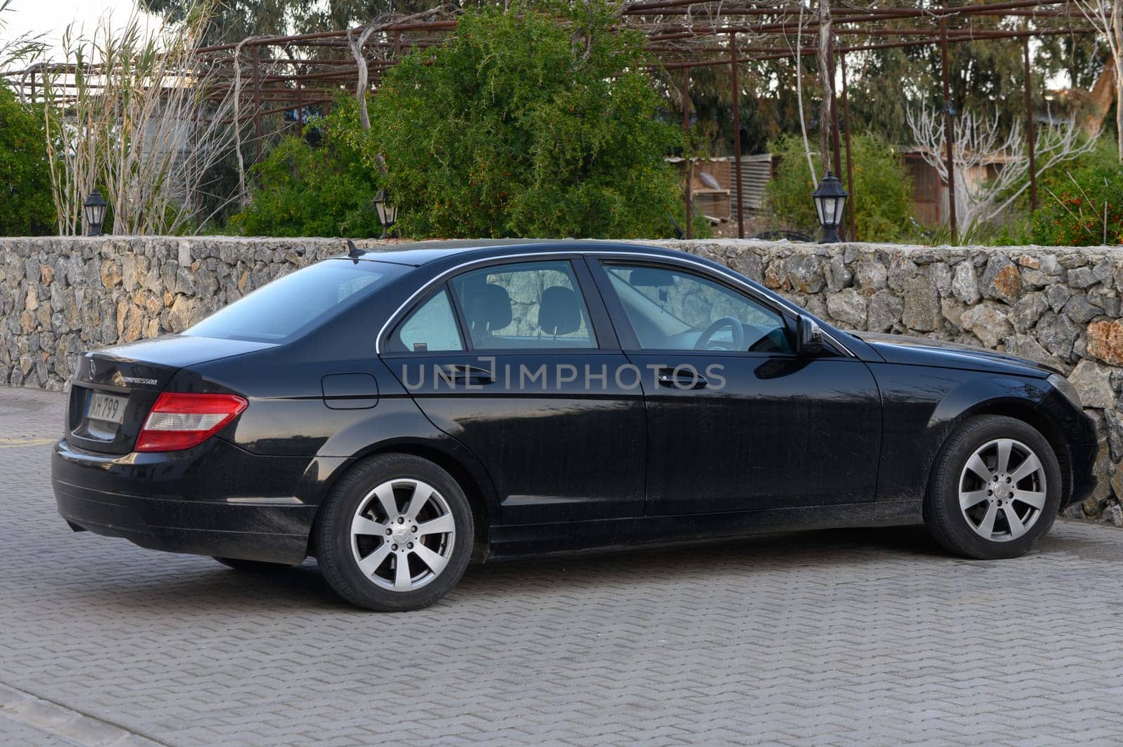 A sleek black sedan rests in a peaceful garden, surrounded by lush greenery and rustic stone walls at sunset.