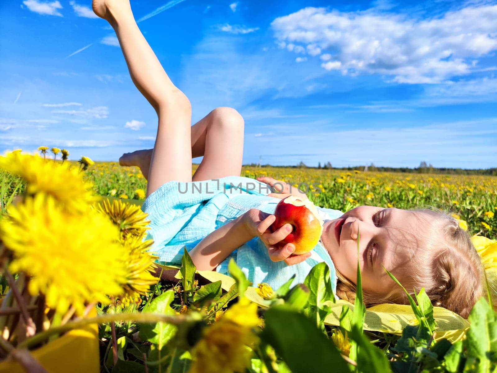 A young girl relaxes in a field of dandelions under a blue sky, happily holding an apple as she stretches out.