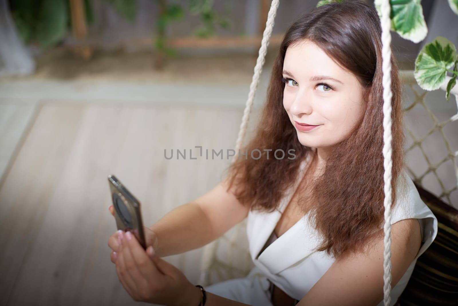 A young woman smiles softly while swinging gently, holding her smartphone in a pleasant indoor space surrounded by plants.