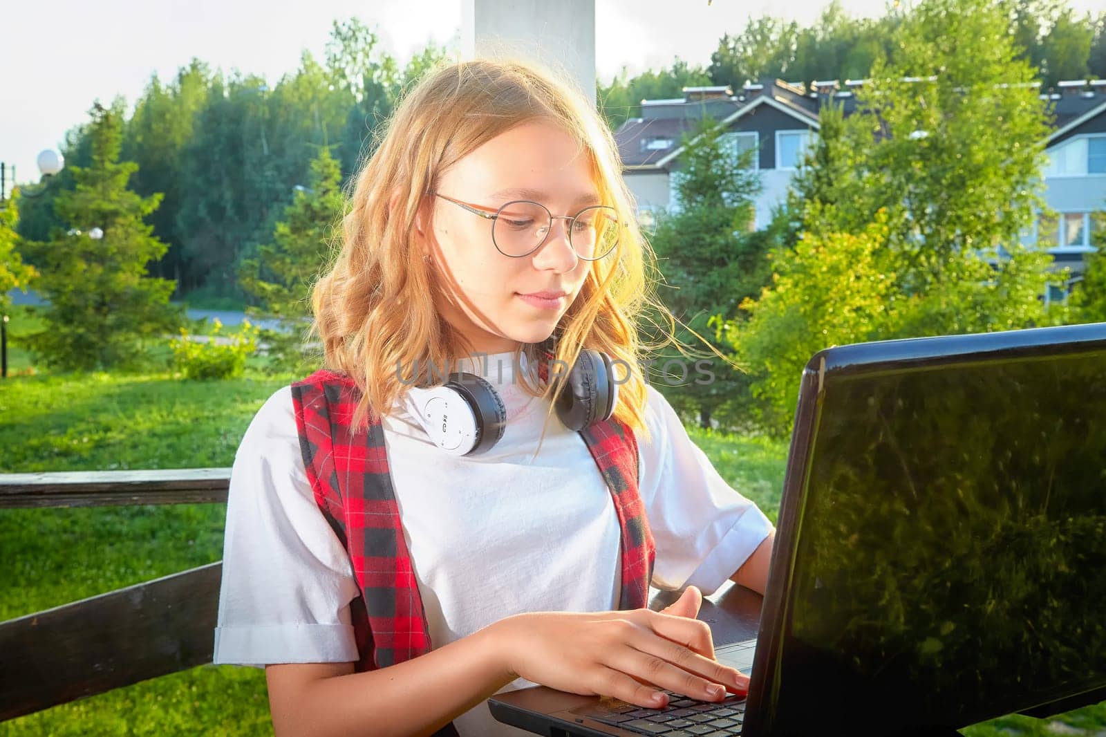 Girl with headphones focuses on laptop screen in a serene outdoor setting, surrounded by greenery on a sunny day.