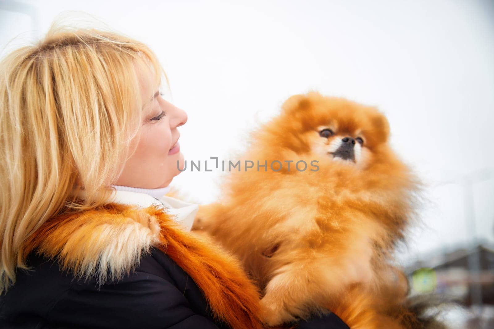 A middle-aged blonde woman holds her fluffy red Pomeranian dog close while walking in the snow on a winter day.