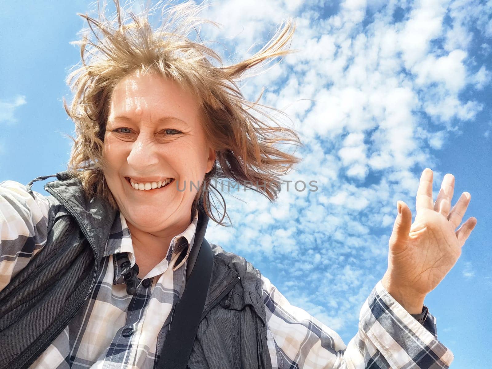 A middle-aged blonde female traveler smiles and waves for a selfie against a beautiful blue sky filled with clouds.