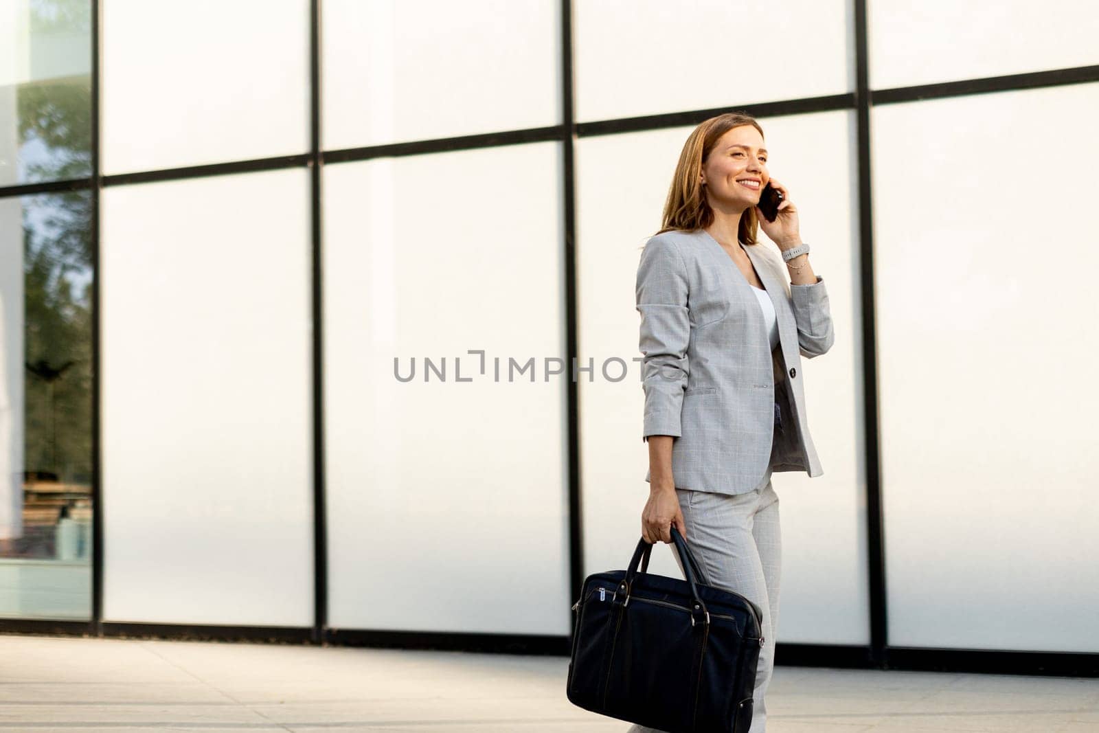Professional woman strolls confidently along a sleek glass structure, her expression joyful as she chats on the phone, dressed in a stylish gray business suit