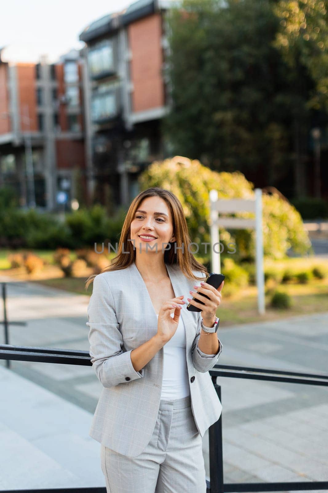 Confident young woman in a light gray suit engages with her smartphone, radiating positivity as she enjoys the lively urban atmosphere filled with greenery